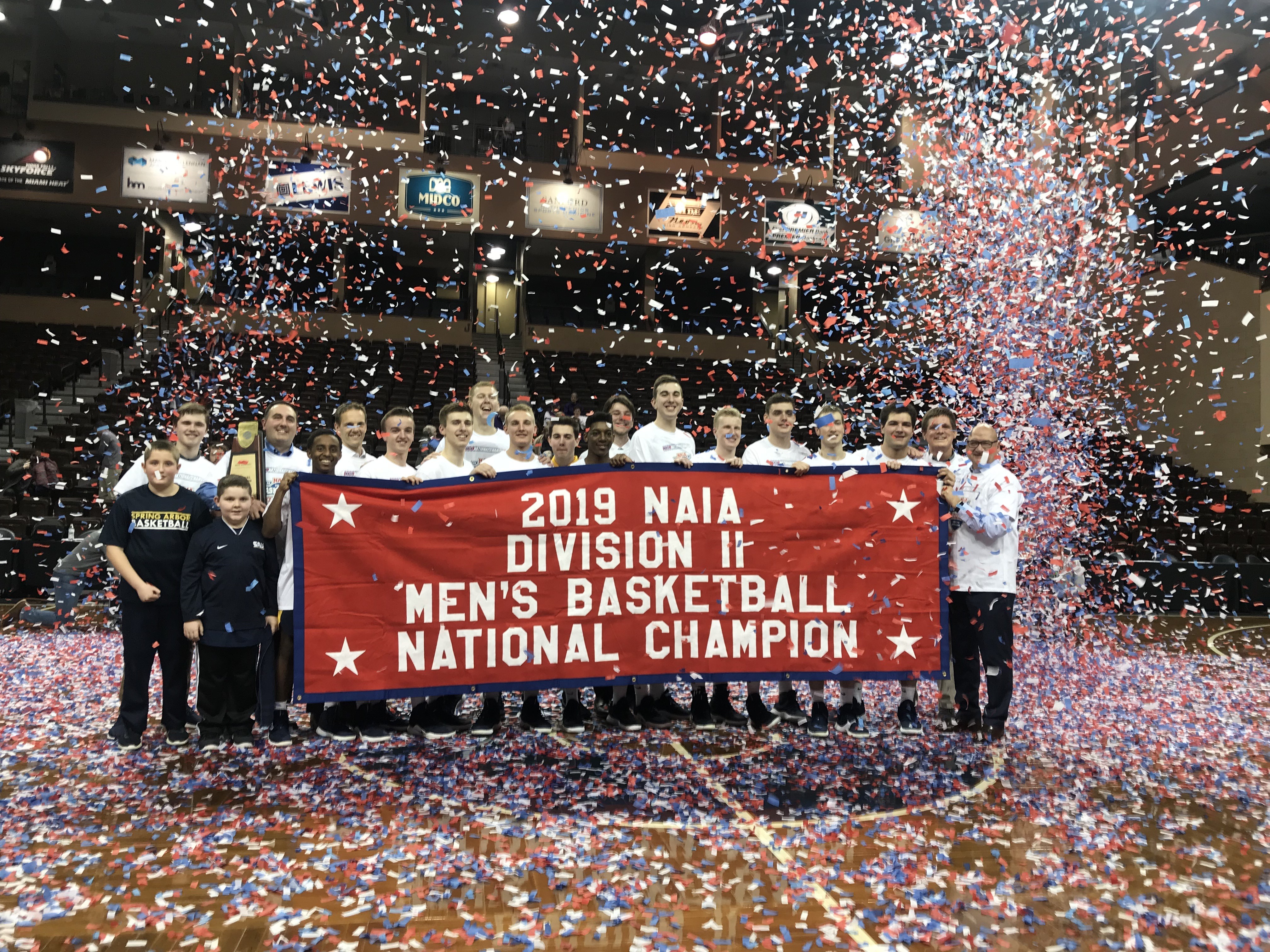 The men's basketball team poses amid confetti behind the NAIA National Champions banner